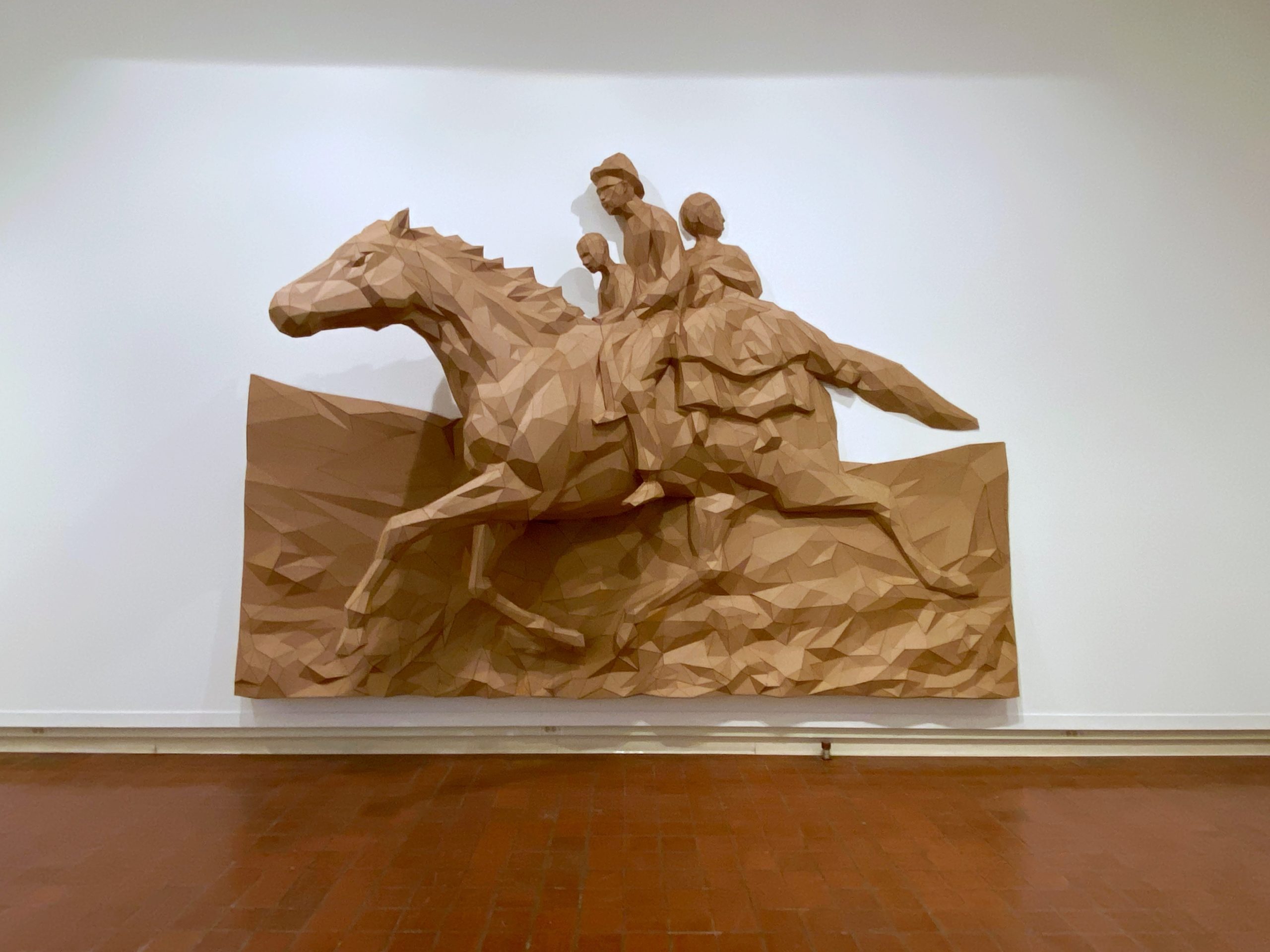 A brown cardboard sculpture, like a relief, of three people riding a horse, mounted on a white gallery wall.