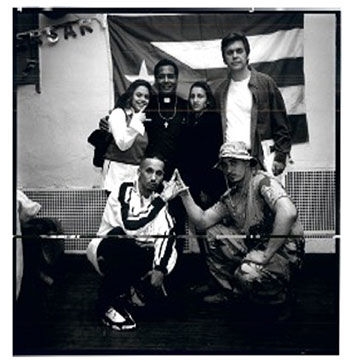 David Brotherton and John Jay professor Dr. Luis Barrios pose in front of a Puerto Rican flag with members of the Latin Kings. The photo is in black and white. Brotherton and Barrios stand at the back with two women, and two men crouch in front, flashing gang hand signs. 