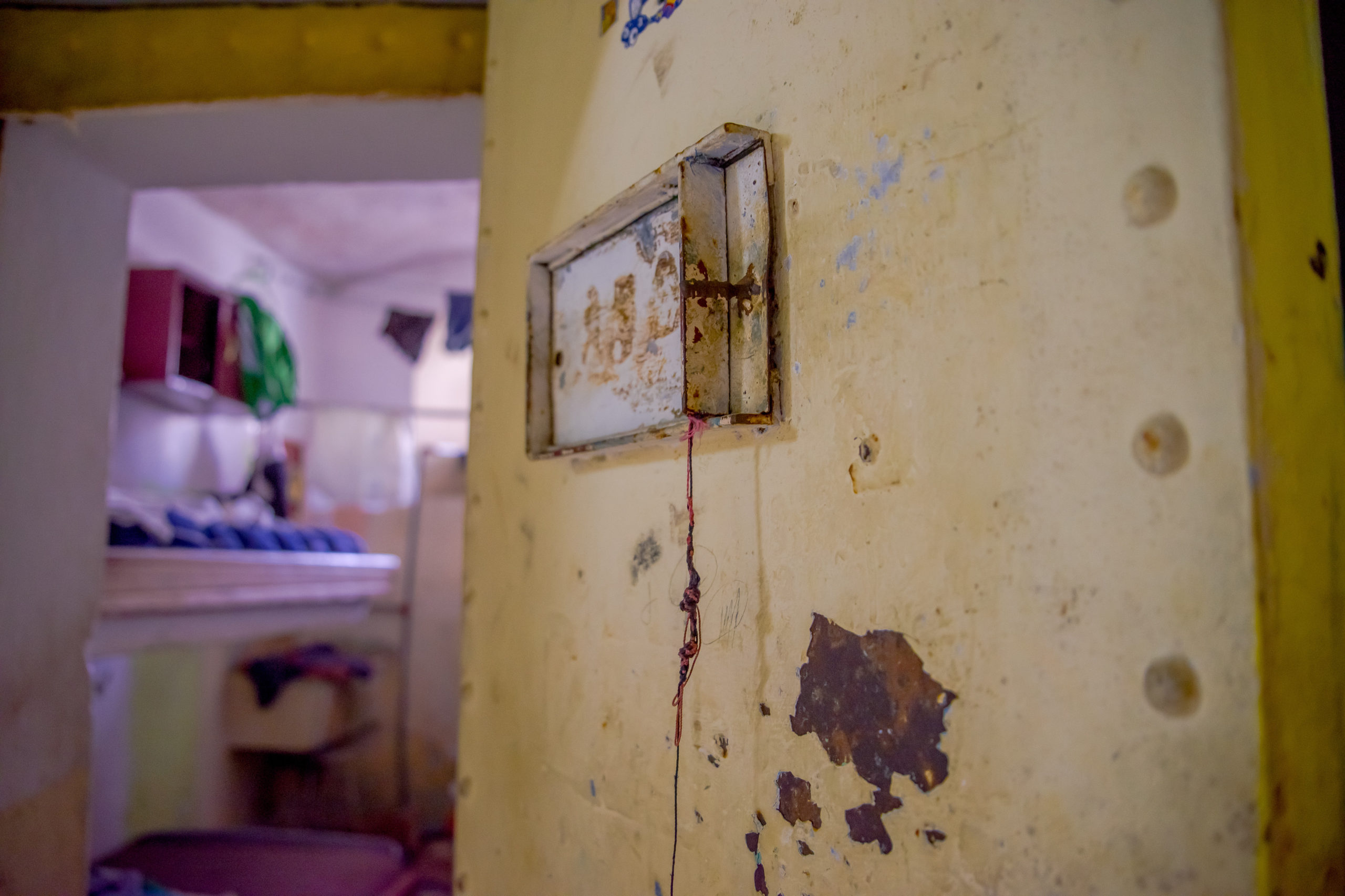 Image shows a grimy yellow wall in the foreground, and an out-of-focus look into a prison cell in the background. 
