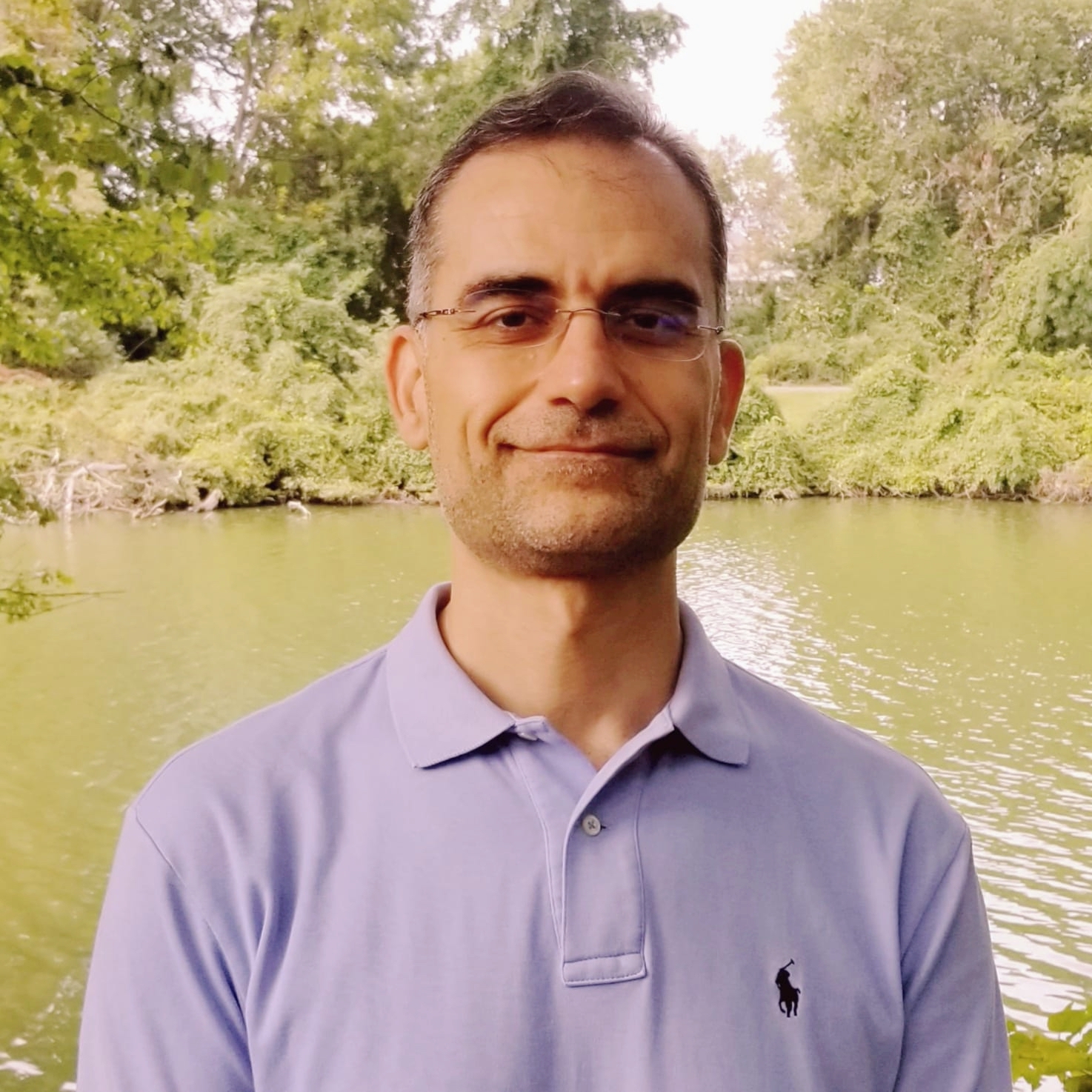 A headshot of Muath Obaidat, looking straight into the camera. He is standing in nature, with a lake and trees behind him, and wearing a purple polo shirt.