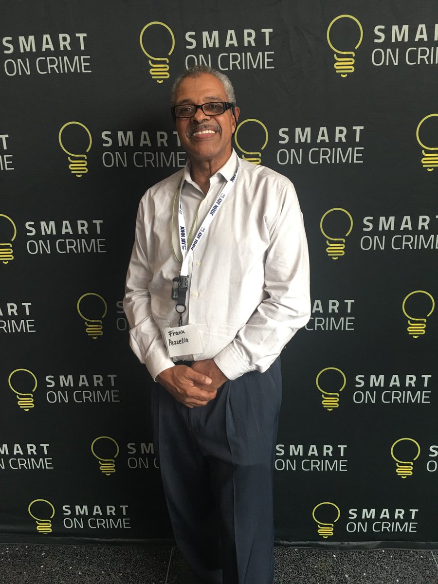 Dr. Frank Pezzella stands in front of a black step and repeat banner that reads "Smart on Crime." He is wearing a white shirt and navy pants, and his hands are folded at his waist. He is smiling into the camera.