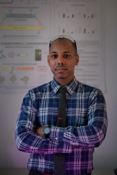 Headshot of Dr. Jason Rauceo. He is facing the camera with his arms folded, glasses on top of his head, wearing a blue plaid shirt and black tie. He is standing in front of a student's poster.