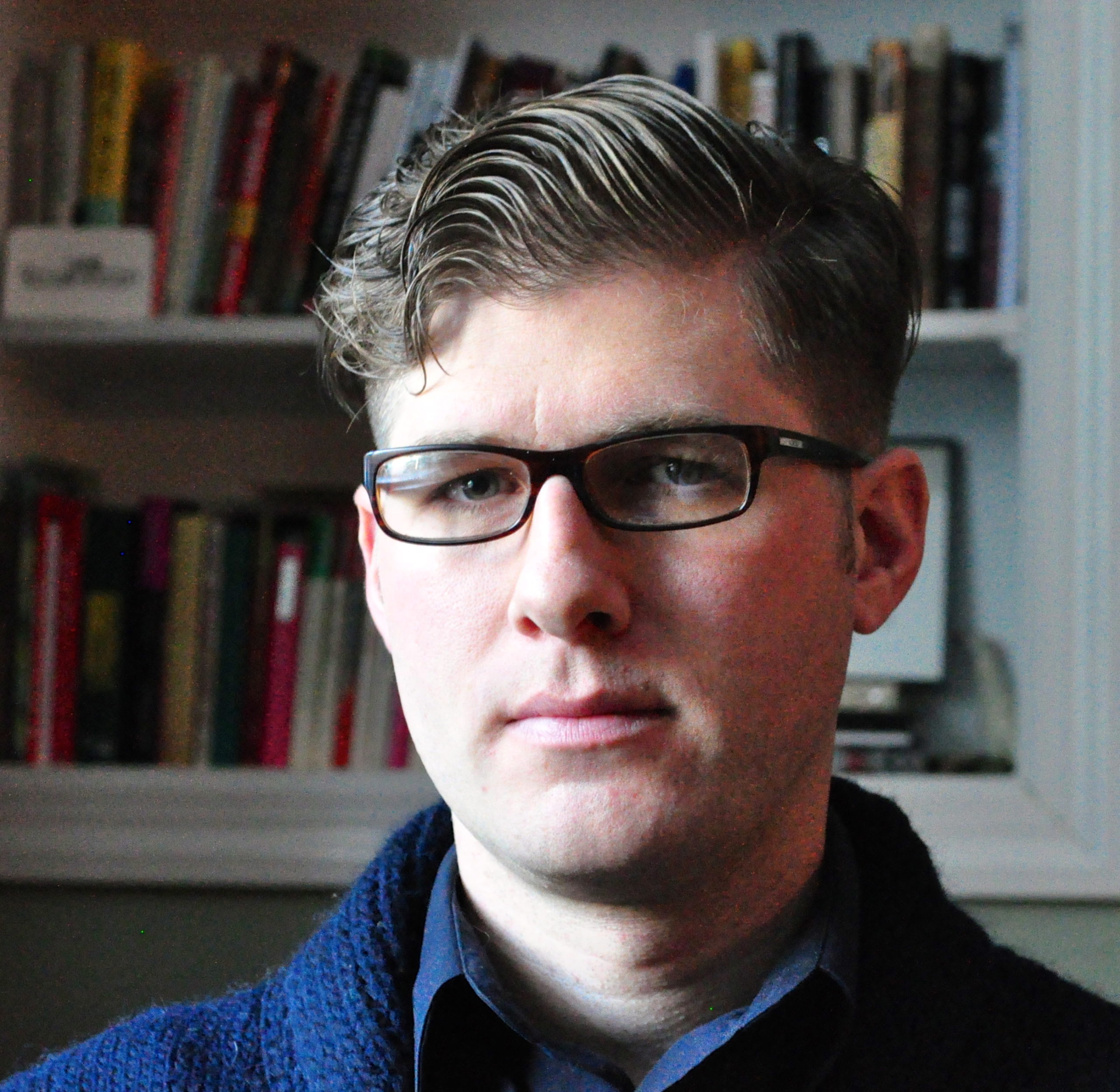 A headshot of Richard Ocejo. He is wearing glasses and a blue shawl collar sweater over a blue shirt, and a bookshelf is in the background. 